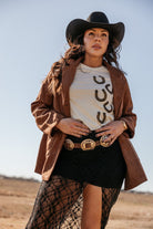 Woman in western boho outfit with black cowboy hat, suede jacket, horseshoe tee, and concho belt