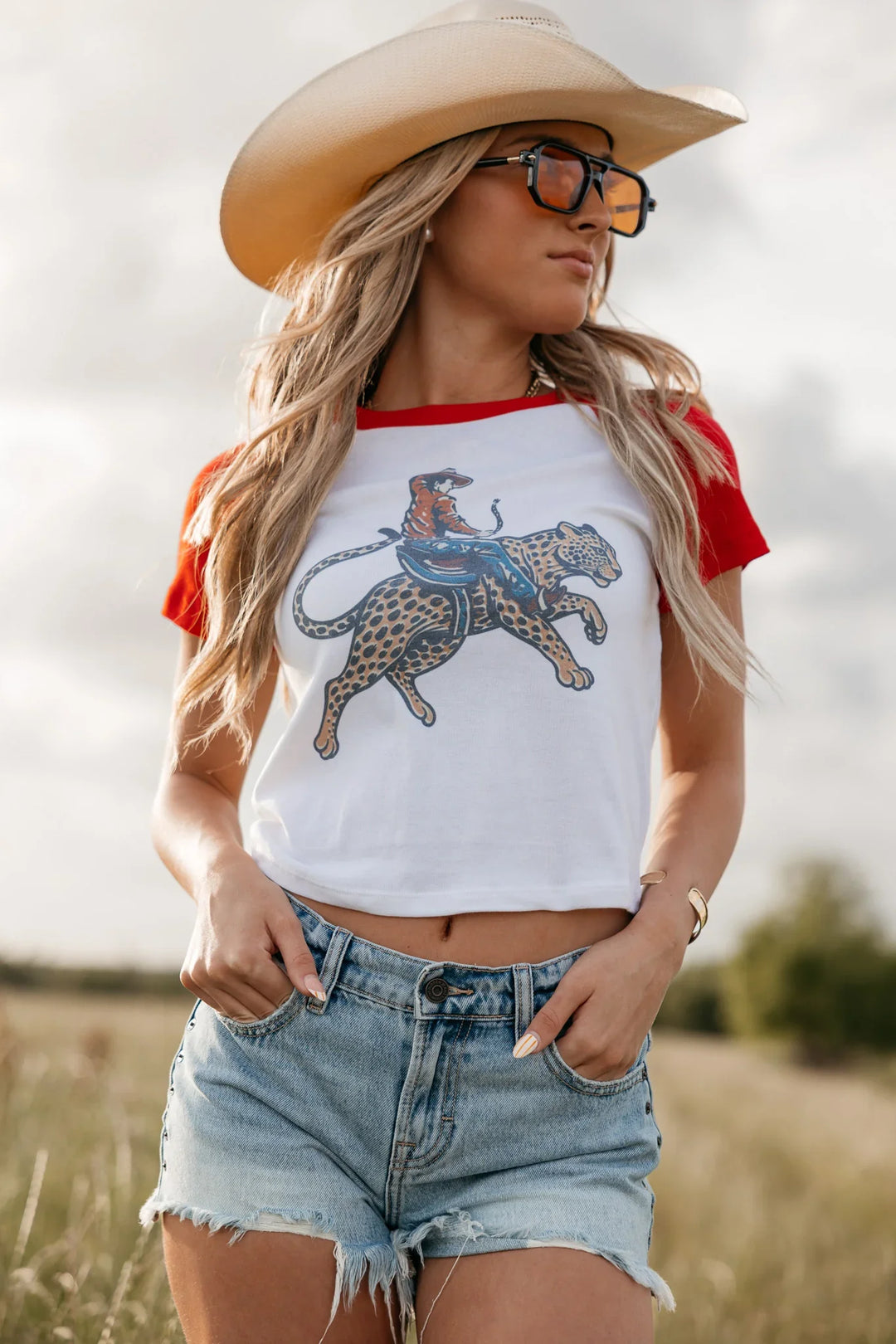 Woman wearing a white and red baseball shirt with a graphic design, denim shorts, and a cowboy hat in a field.