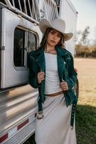 Woman in a suede teal western jacket, white skirt, and cowgirl hat by a horse trailer outdoors