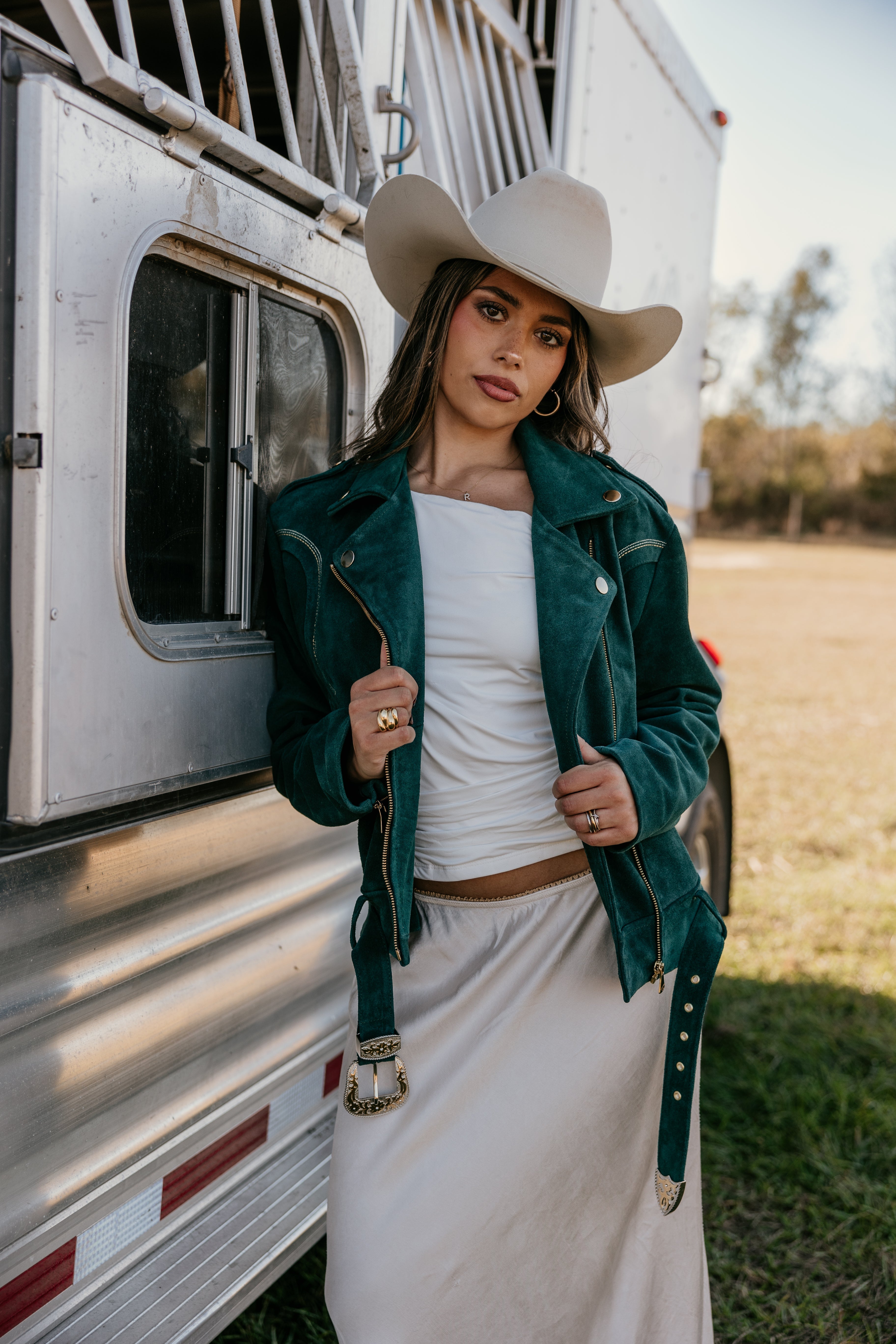 Woman in a suede teal western jacket, white skirt, and cowgirl hat by a horse trailer outdoors