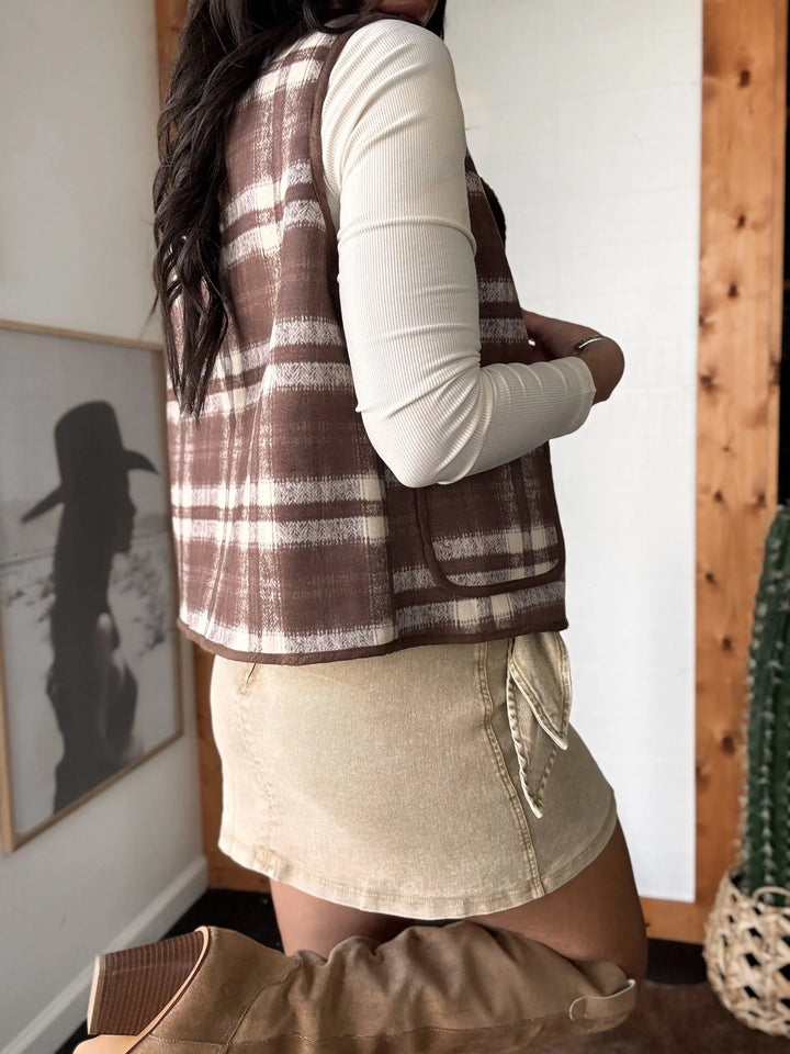 Person wearing a plaid vest over a white long-sleeve shirt and beige skirt in a room with wooden walls.