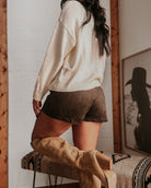 Woman in cream sweater, brown suede shorts, and western knee-high boots, rustic decor