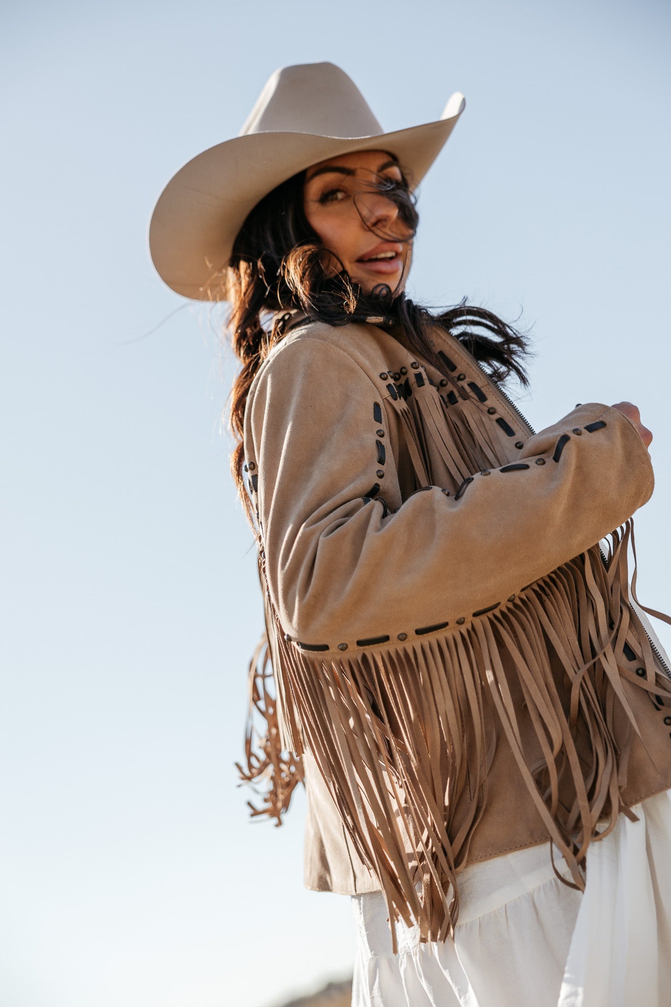 Woman in tan fringe western jacket, white dress, and cowgirl hat outdoors