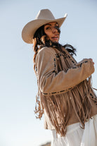 Woman in tan fringe western jacket, white dress, and cowgirl hat outdoors