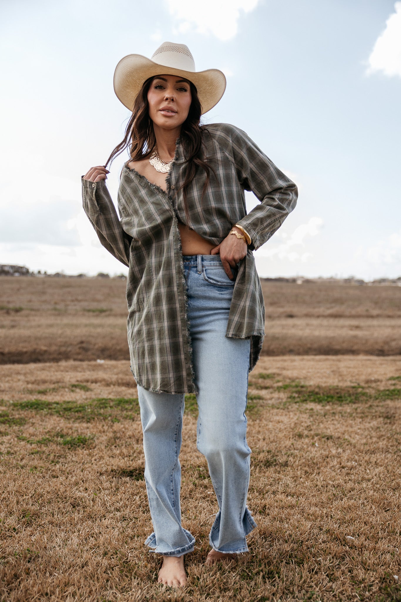 Woman in a plaid shirt, cowboy hat, and light wash jeans standing in a western field