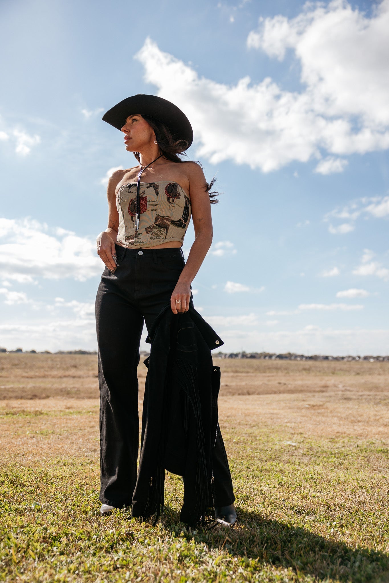 Woman in western boho outfit with black cowboy hat, patterned tube top, and flared pants outdoors.
