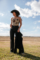 Woman in western boho outfit with black cowboy hat, patterned tube top, and flared pants outdoors.