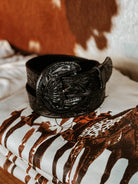 Tooled black western belt with ornate buckle on folded cowgirl graphic tee, cow print background