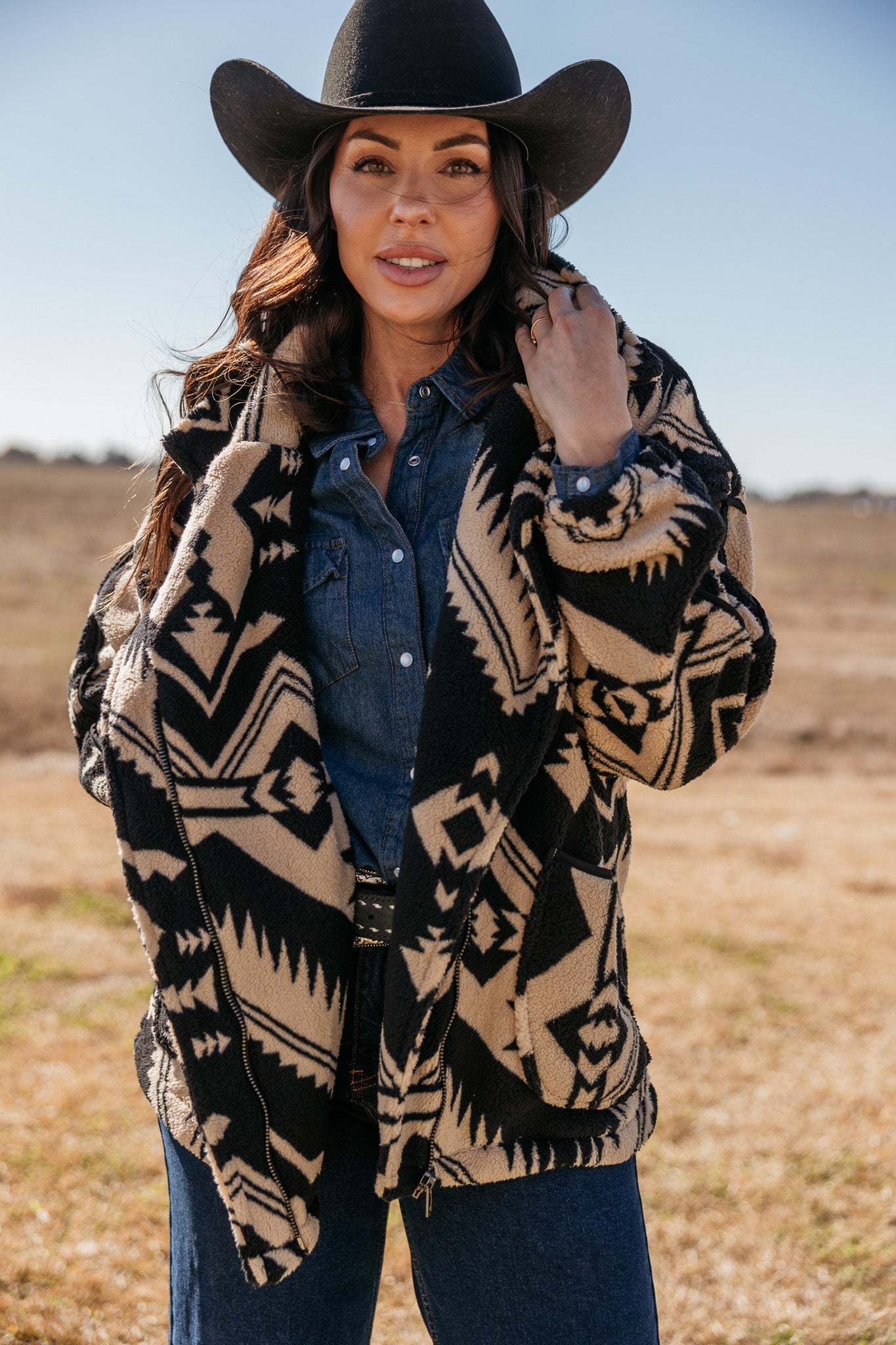 Woman in western boho outfit with black cowboy hat, denim shirt, and Aztec print jacket outdoors