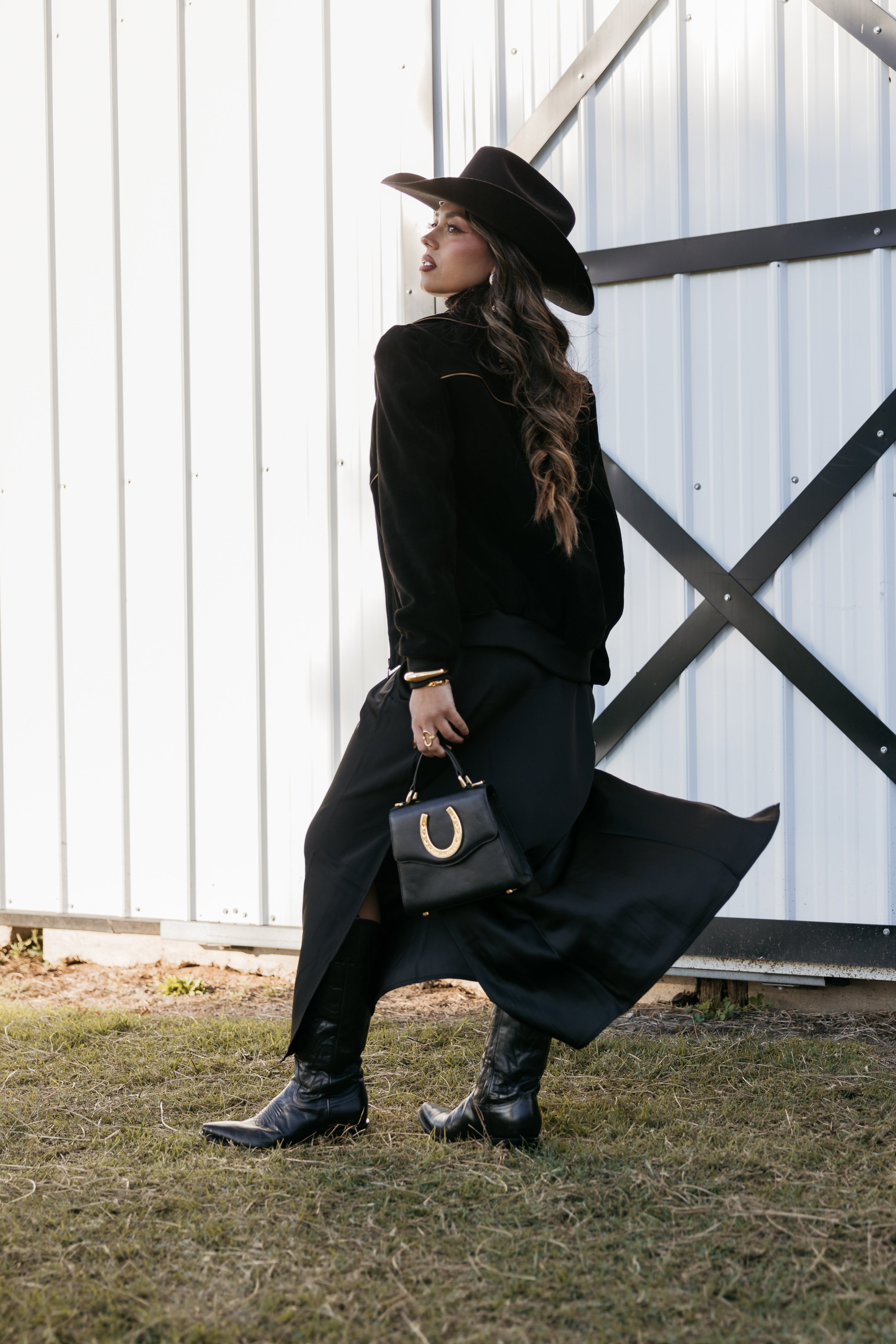 Woman in black cowgirl hat and western outfit with boots and horseshoe handbag outdoors