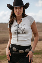Woman in cowgirl hat, 'Life is a Rodeo' graphic tee, concho belt, and western jewelry outdoors