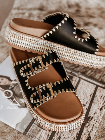 Black platform sandals featuring two stitched buckle straps with gold adjustable hardware, a braided raffia-style platform, textured sole for added grip, and a soft cushioned insole for comfort.