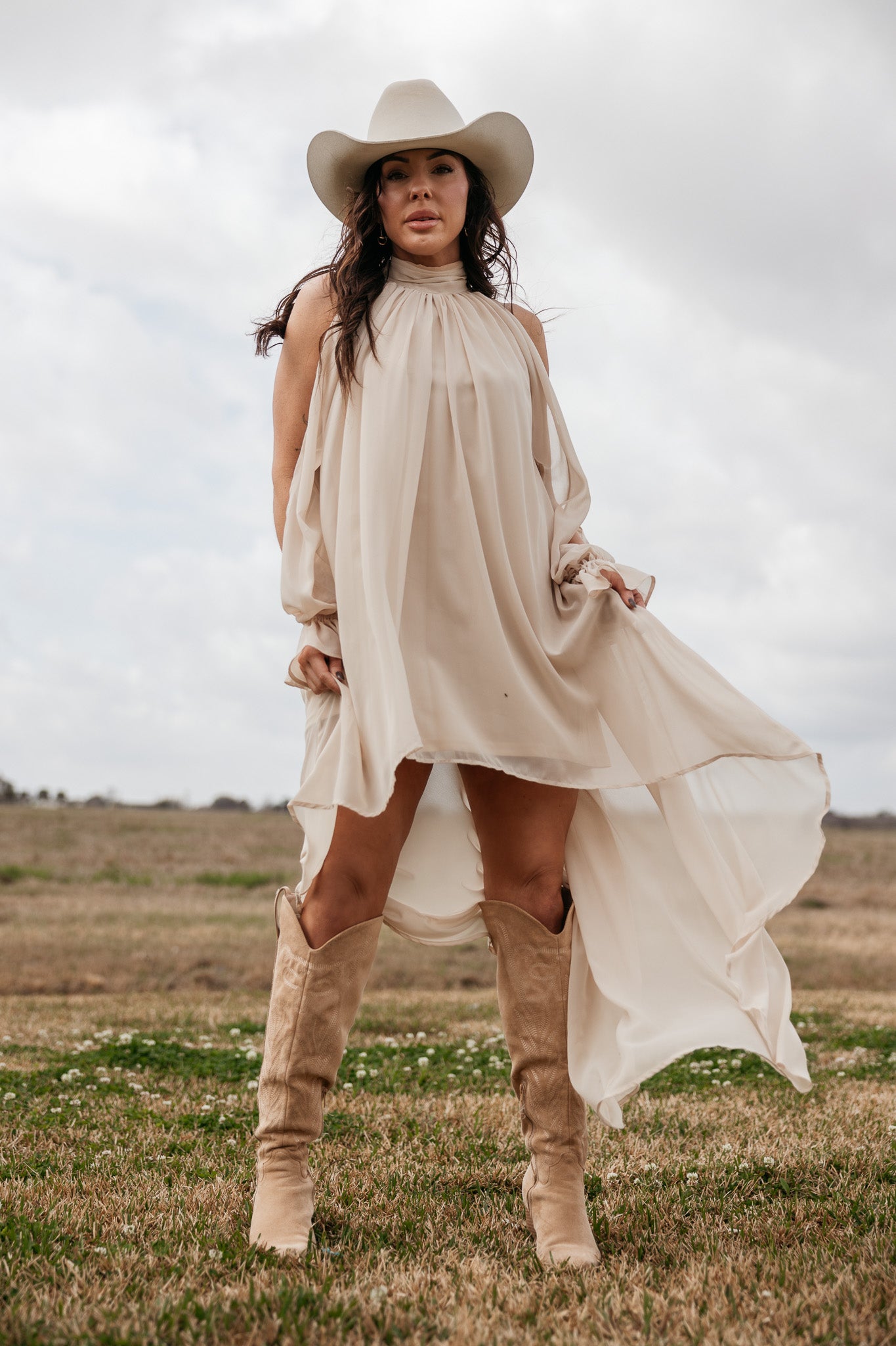 Woman in cream western boho dress, cowboy hat, and boots standing in open field