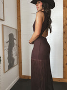 Woman wearing a long brown dress with fringe details, standing in a room with wooden walls and framed pictures.