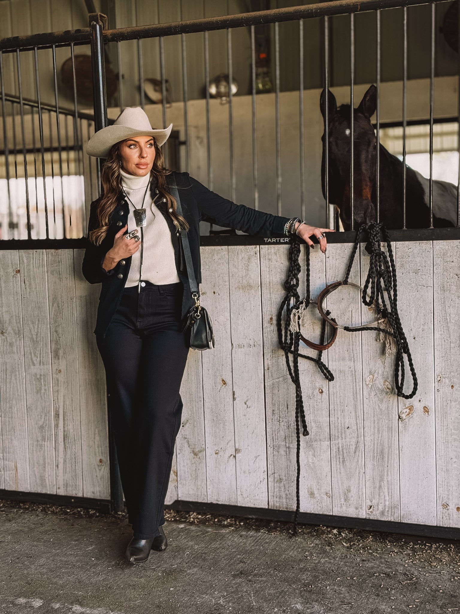 Woman in western outfit with cowboy hat poses by horse stable, showcasing cowgirl style fashion.