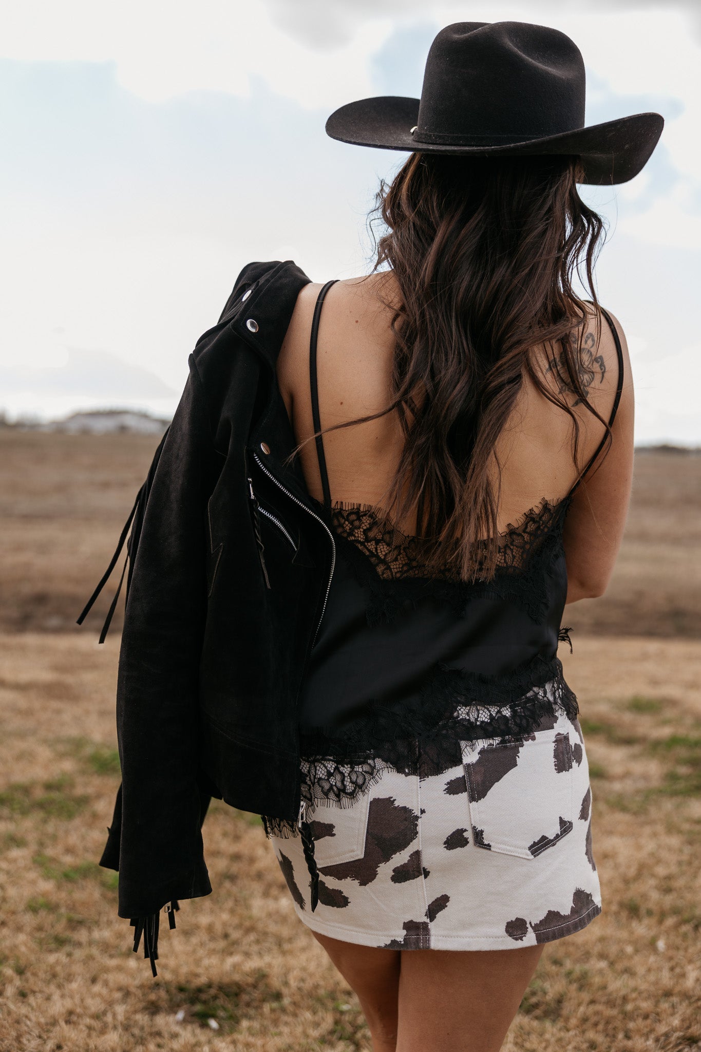 Woman in black cowboy hat, black lace cami, and cow print skirt with suede jacket outdoors