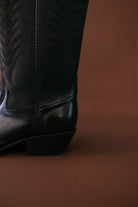 Black leather western cowgirl boot with stitched patterns, close-up on brown background
