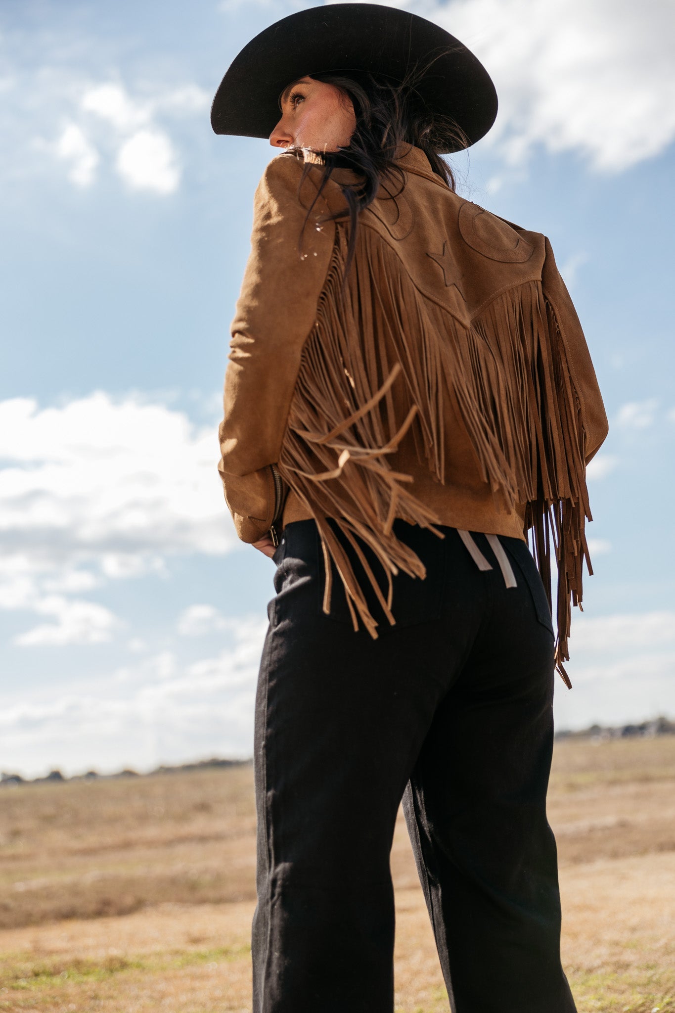 Woman in brown suede fringe jacket and black cowboy hat outdoors, western boho fashion