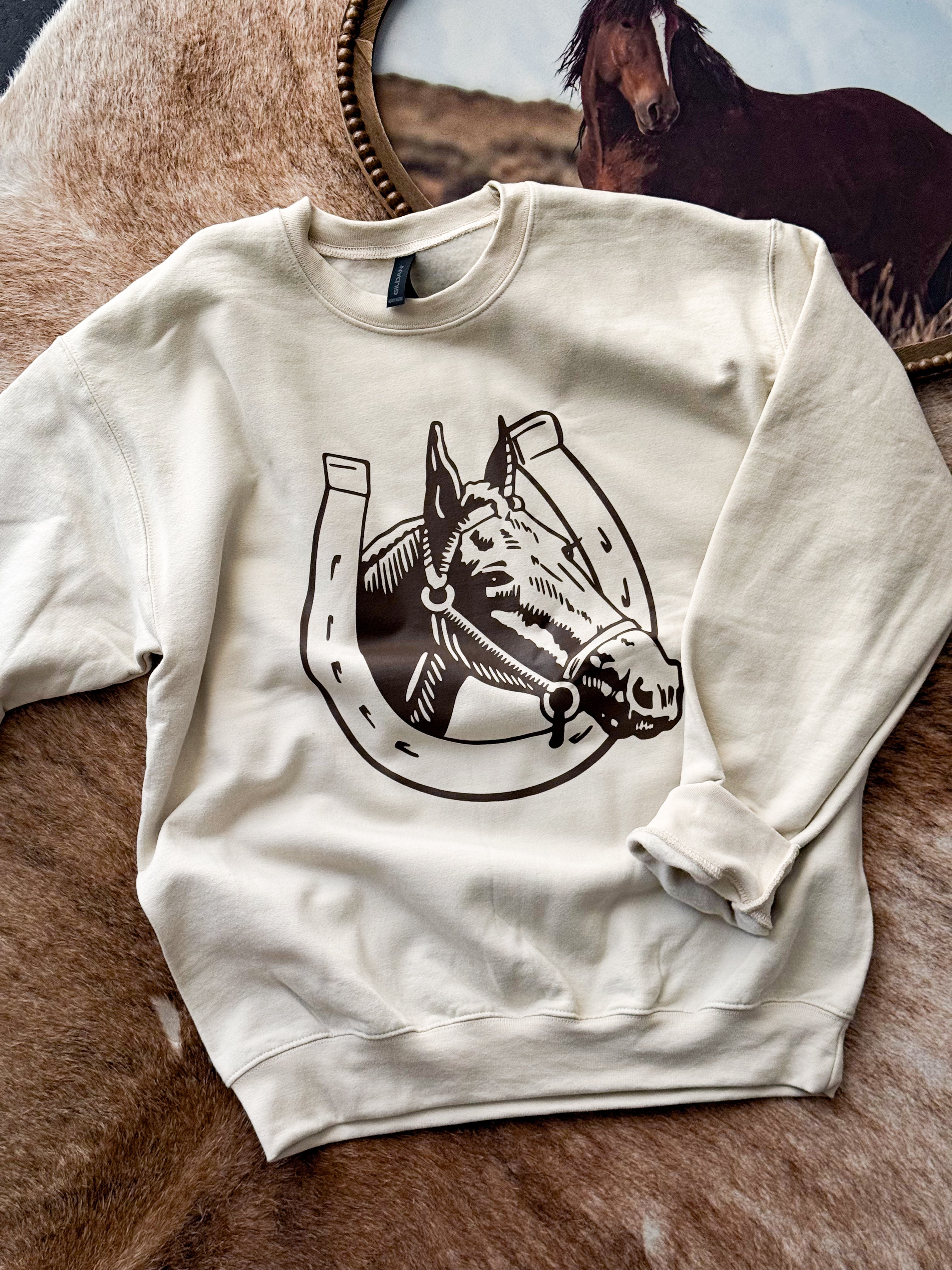 Cream western sweatshirt with horse and horseshoe graphic, laid on cowhide rug
