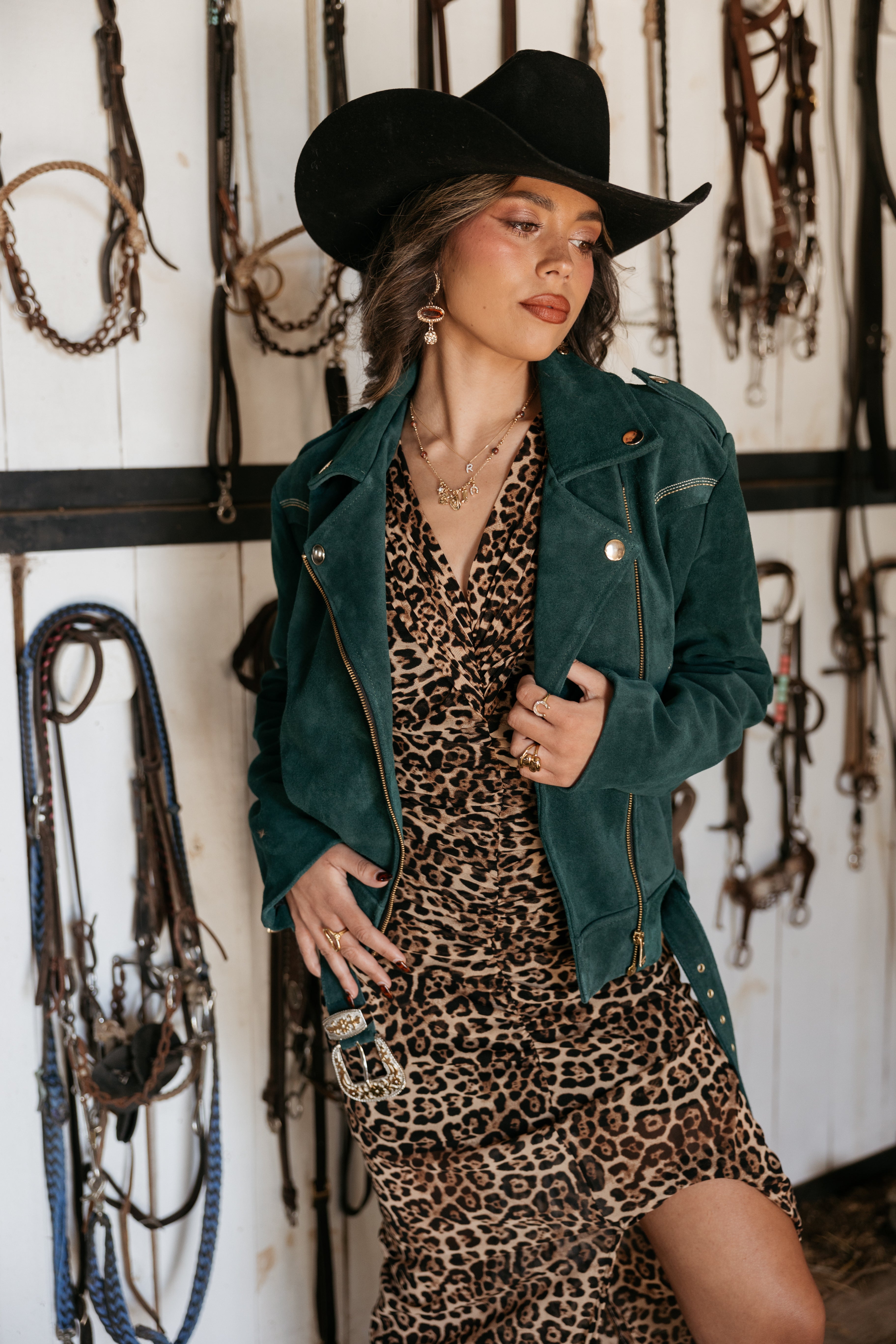 Woman in black cowboy hat, green suede jacket, and leopard print western dress with layered jewelry, standing in a tack room.