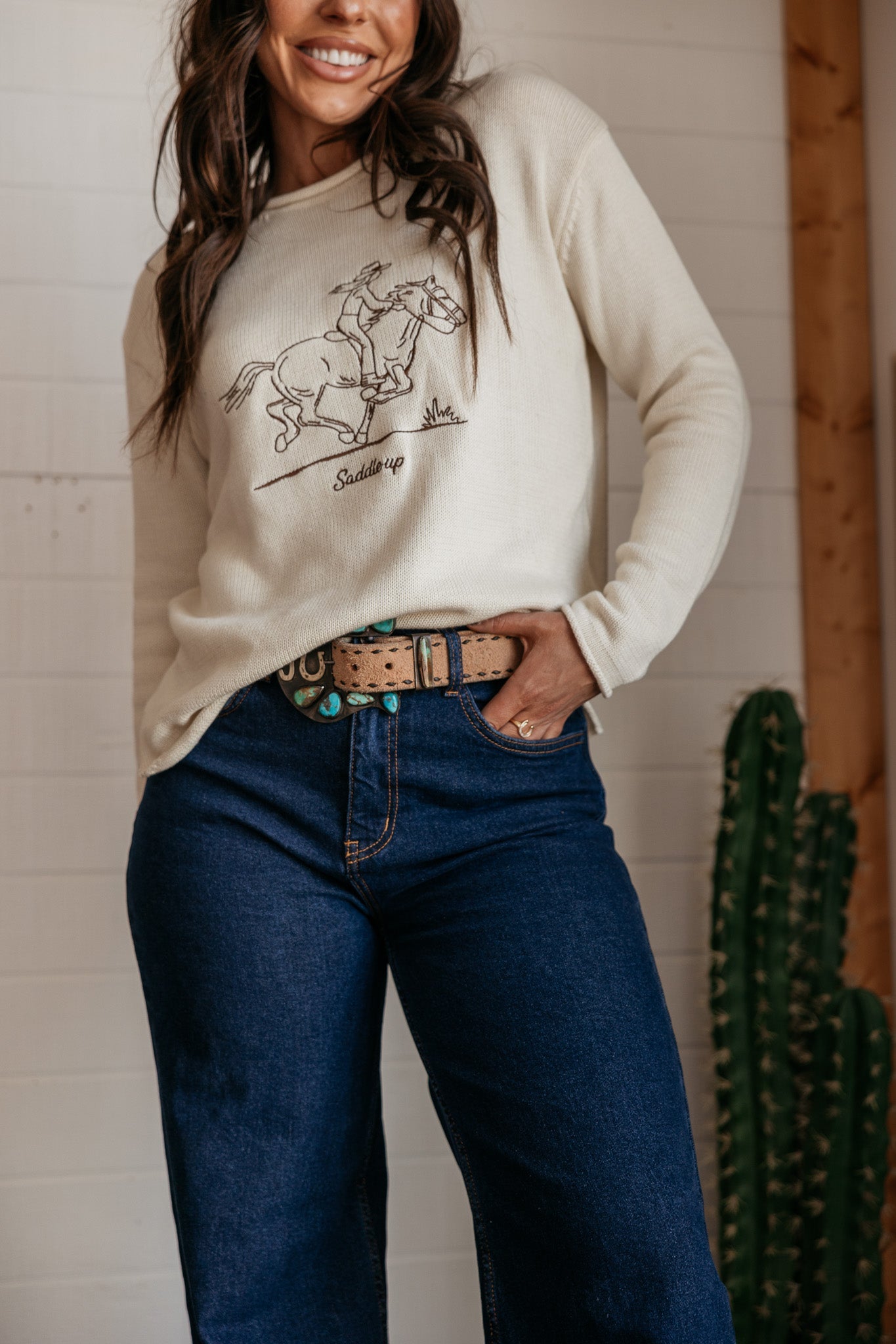 Woman in western graphic sweater, turquoise belt, and high-waisted jeans standing indoors