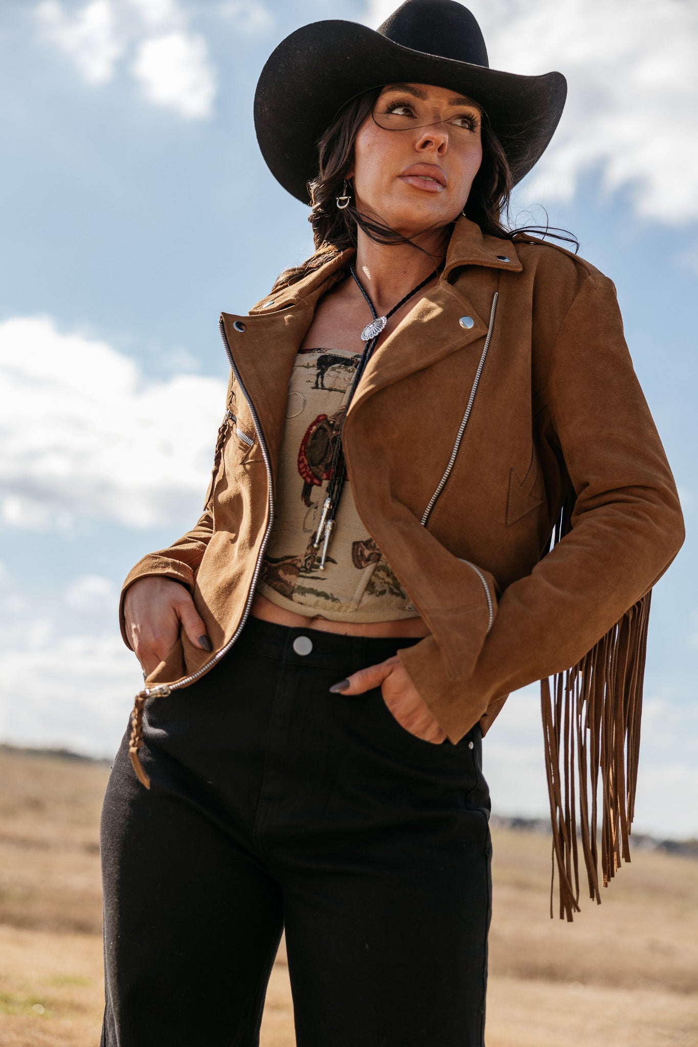 Woman in a brown fringe suede jacket, western crop top, black jeans, and cowboy hat outdoors