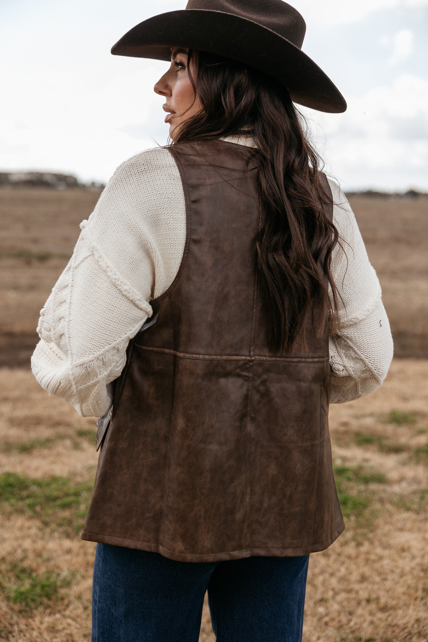 Woman in brown felt cowboy hat and brown leather vest with cream knit sweater, outdoors