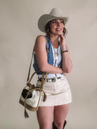 Smiling woman in cowboy hat, denim vest, cream mini skirt, Western bag and jewelry.