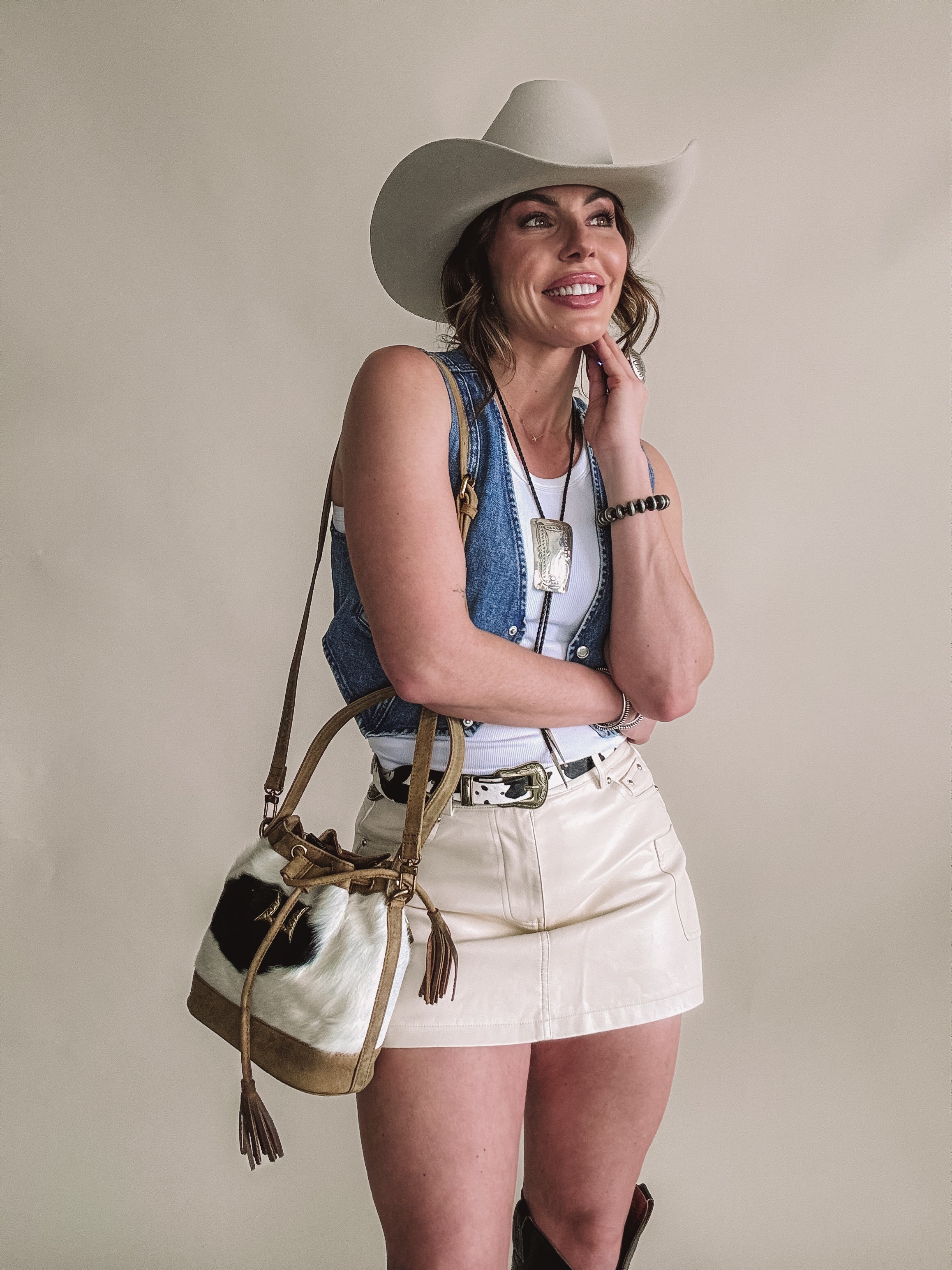 Smiling woman in cowboy hat, denim vest, cream mini skirt, Western bag and jewelry.