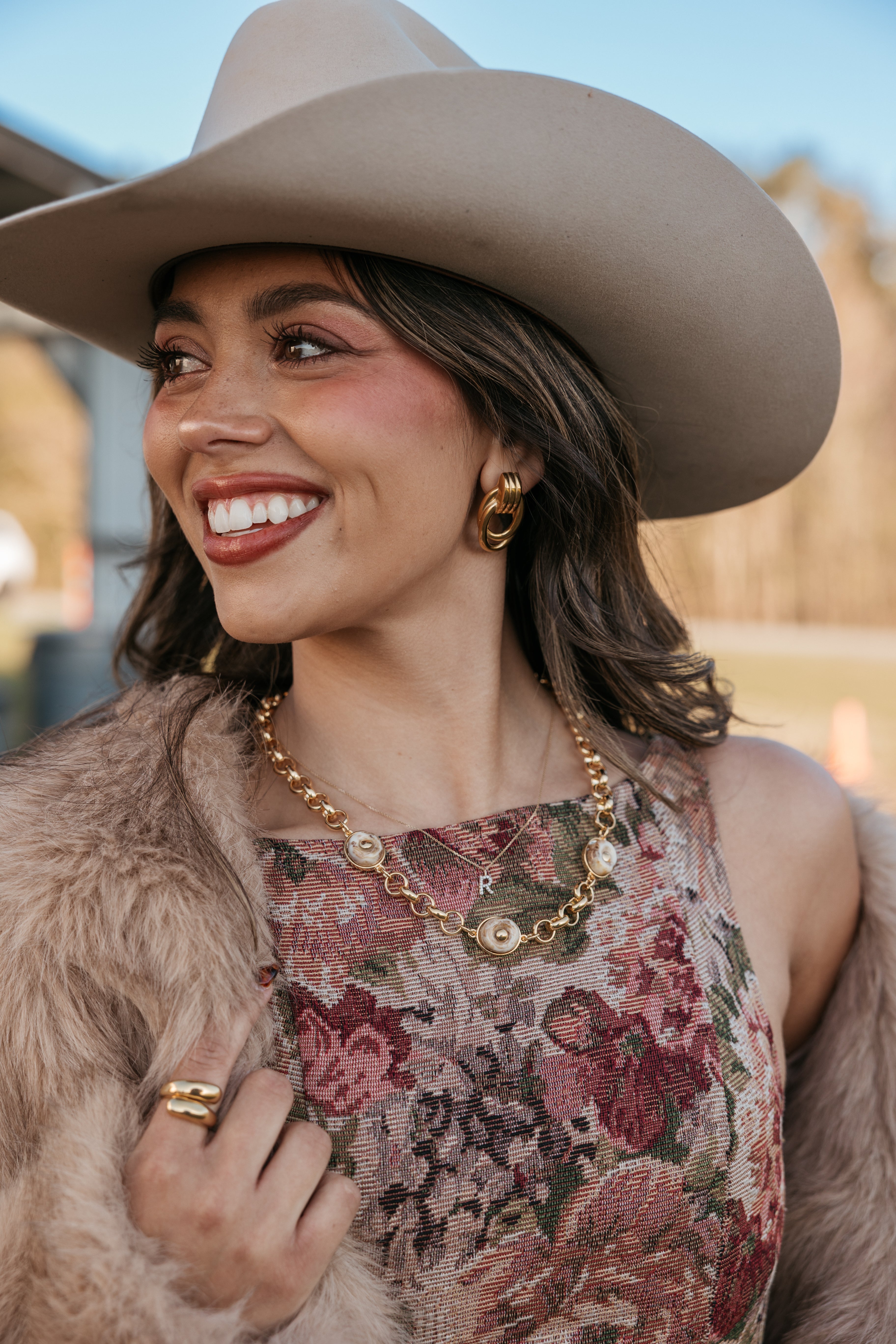 Woman in cowgirl hat, floral western dress, gold jewelry, and faux fur jacket outdoors
