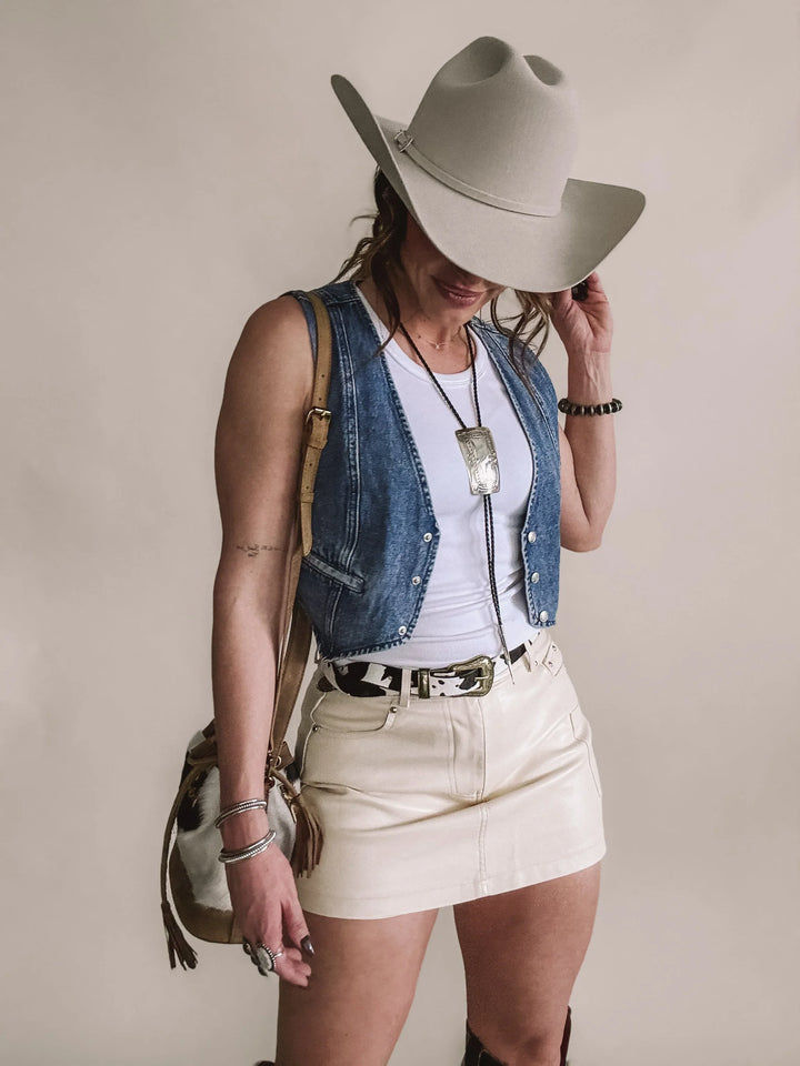 Woman in a western cowgirl outfit with felt hat, denim vest, cream mini skirt, and cow print belt