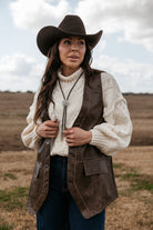 Woman in western hat, cream sweater, brown vest, and bolo tie standing outdoors
