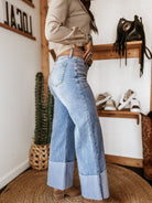 Woman wearing western boho wide-leg cuffed jeans, cowgirl boots, and a plaid shirt in a rustic room.