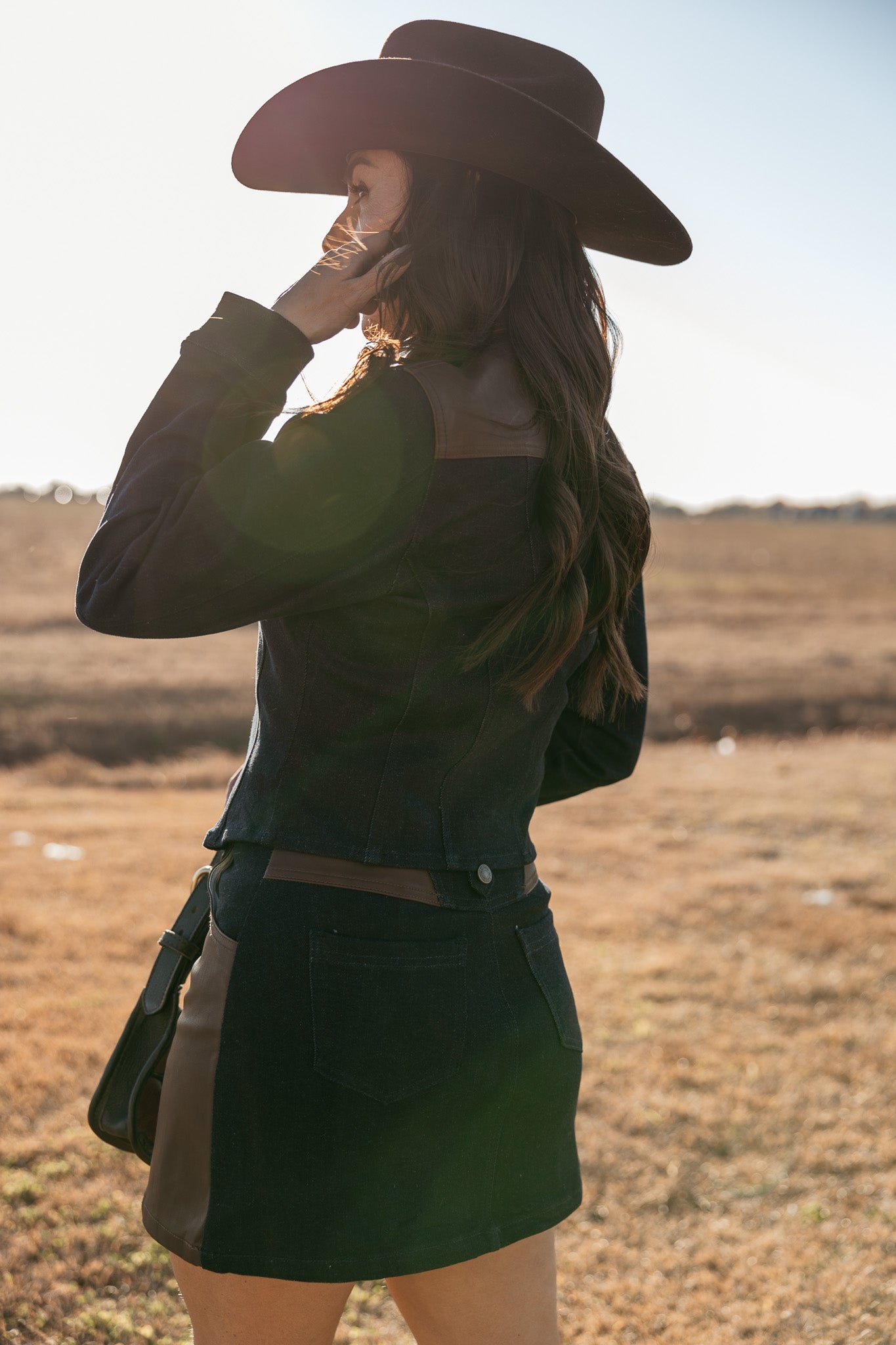 Woman in western denim jacket, mini skirt, and black cowboy hat outdoors