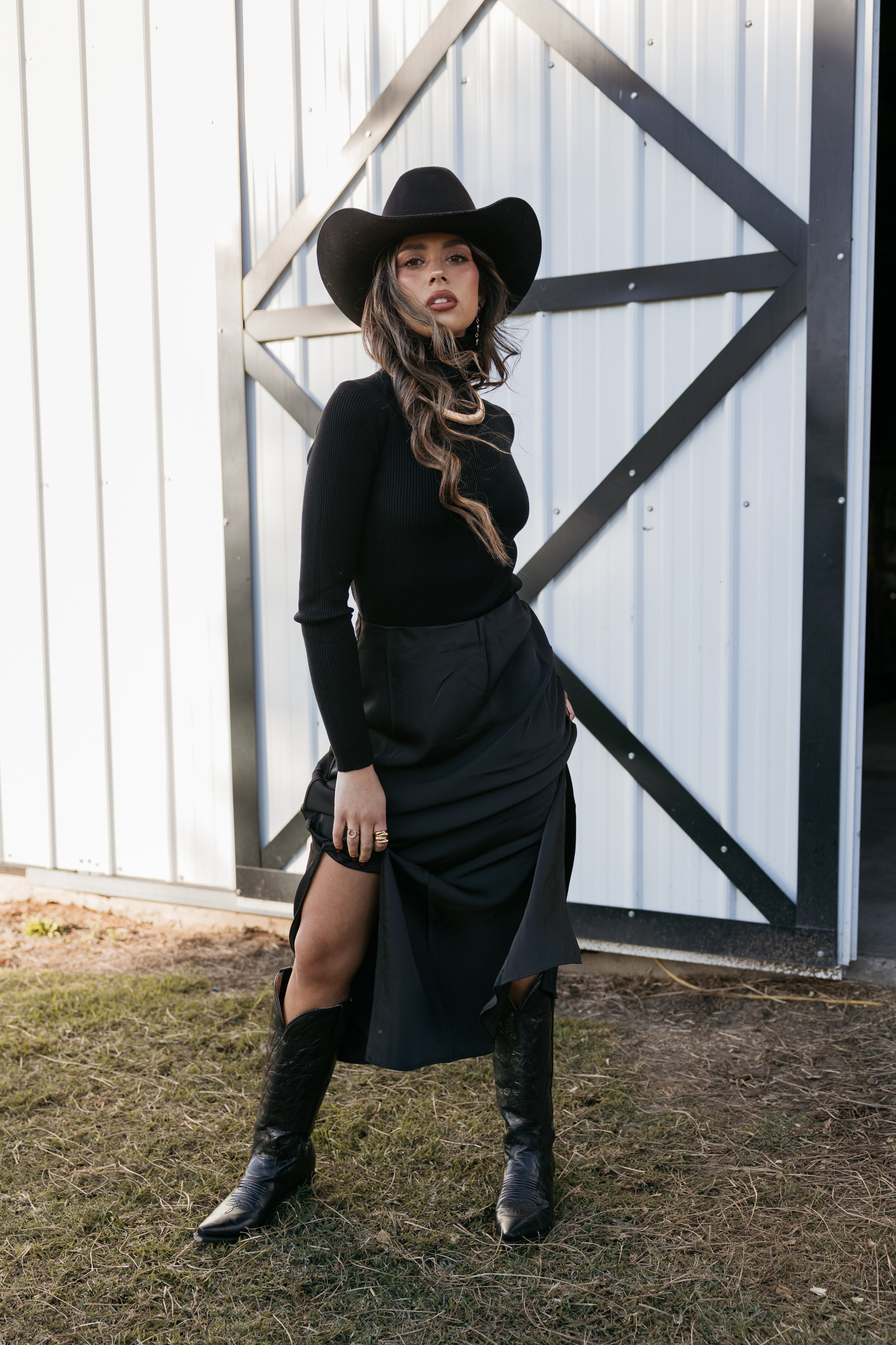 Woman in black cowboy hat, black long sleeve top, skirt, and boots posing in front of barn