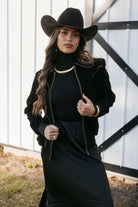 Woman in black cowgirl hat, western boho jacket, and jewelry posing by barn door