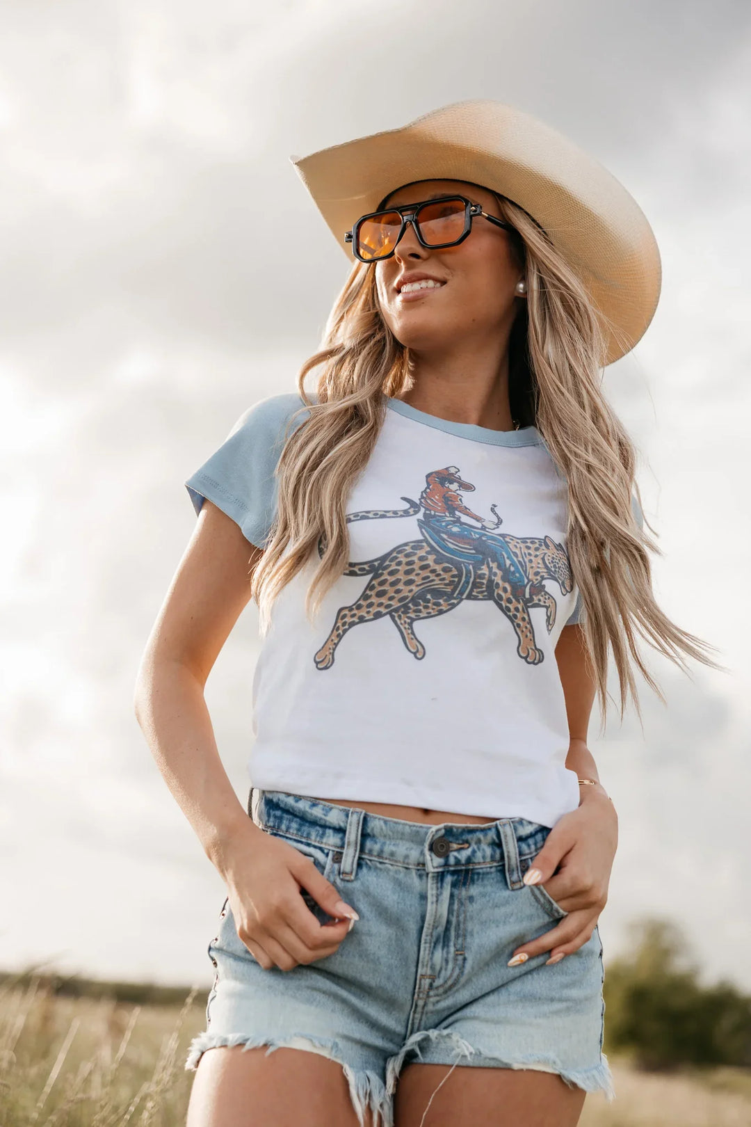 Woman wearing a graphic t-shirt with a horse and leopard design, denim shorts, and a wide-brimmed hat outdoors.