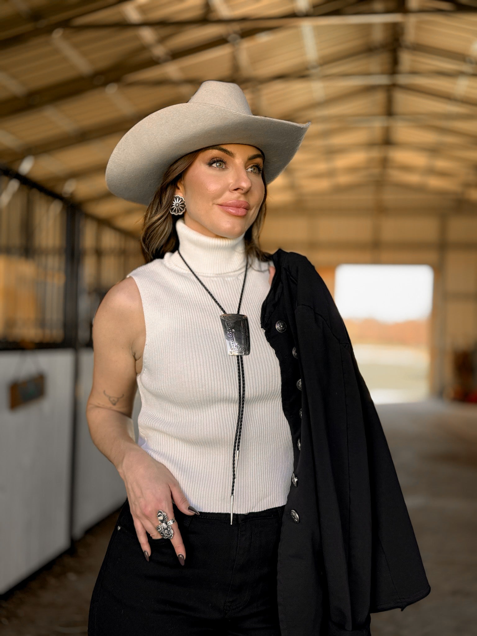 Woman in a white cowboy hat, sleeveless turtleneck, bolo tie, and western jewelry in a barn.