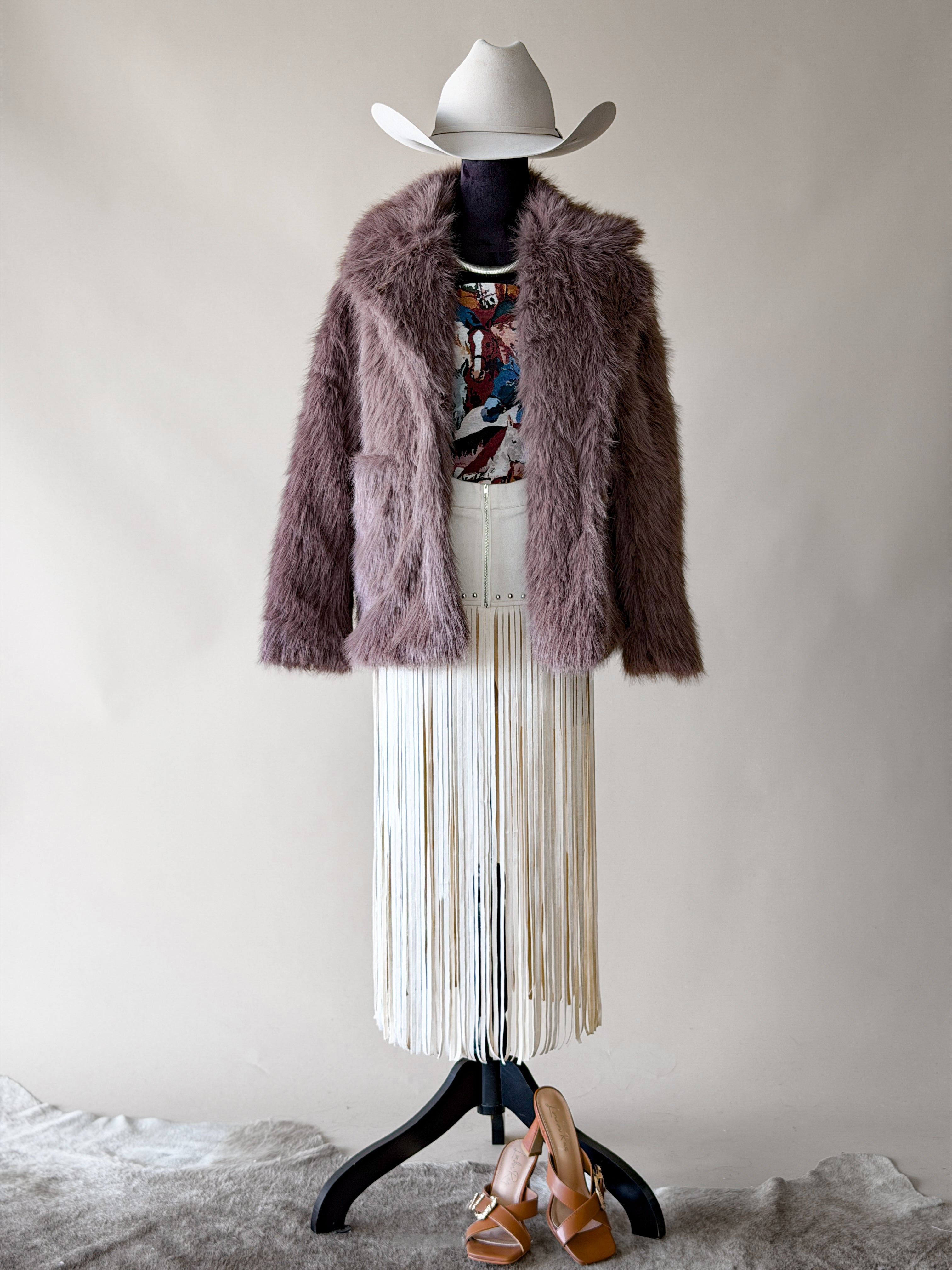 Western boho outfit with white cowboy hat, faux fur jacket, graphic tee, fringe skirt, sandals