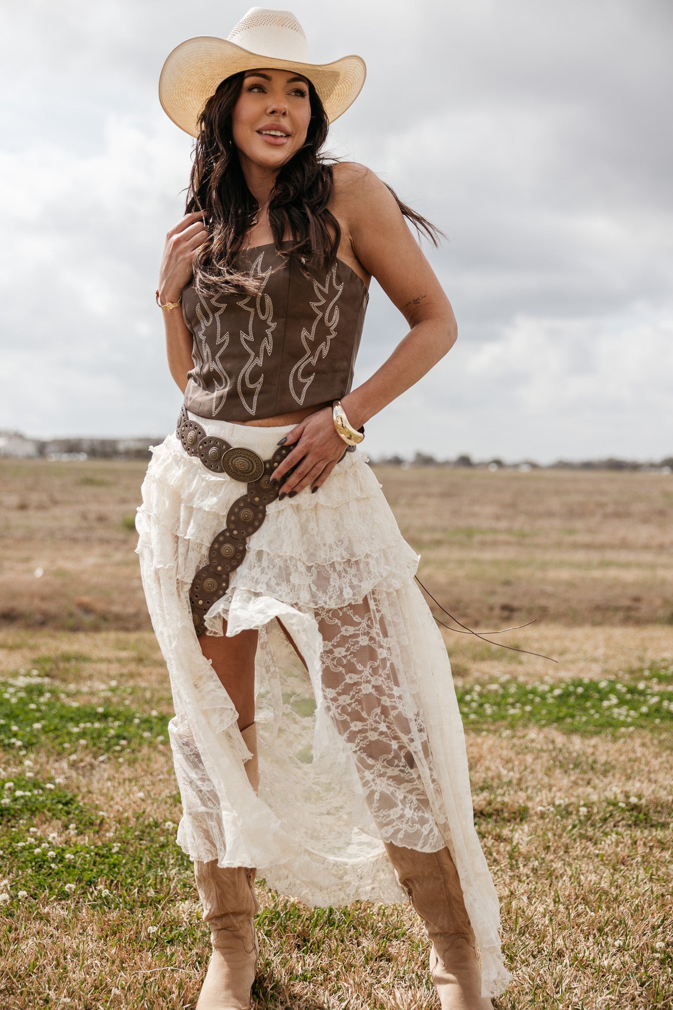 Woman in cowgirl hat, brown corset top, lace western skirt, and boots standing in a field
