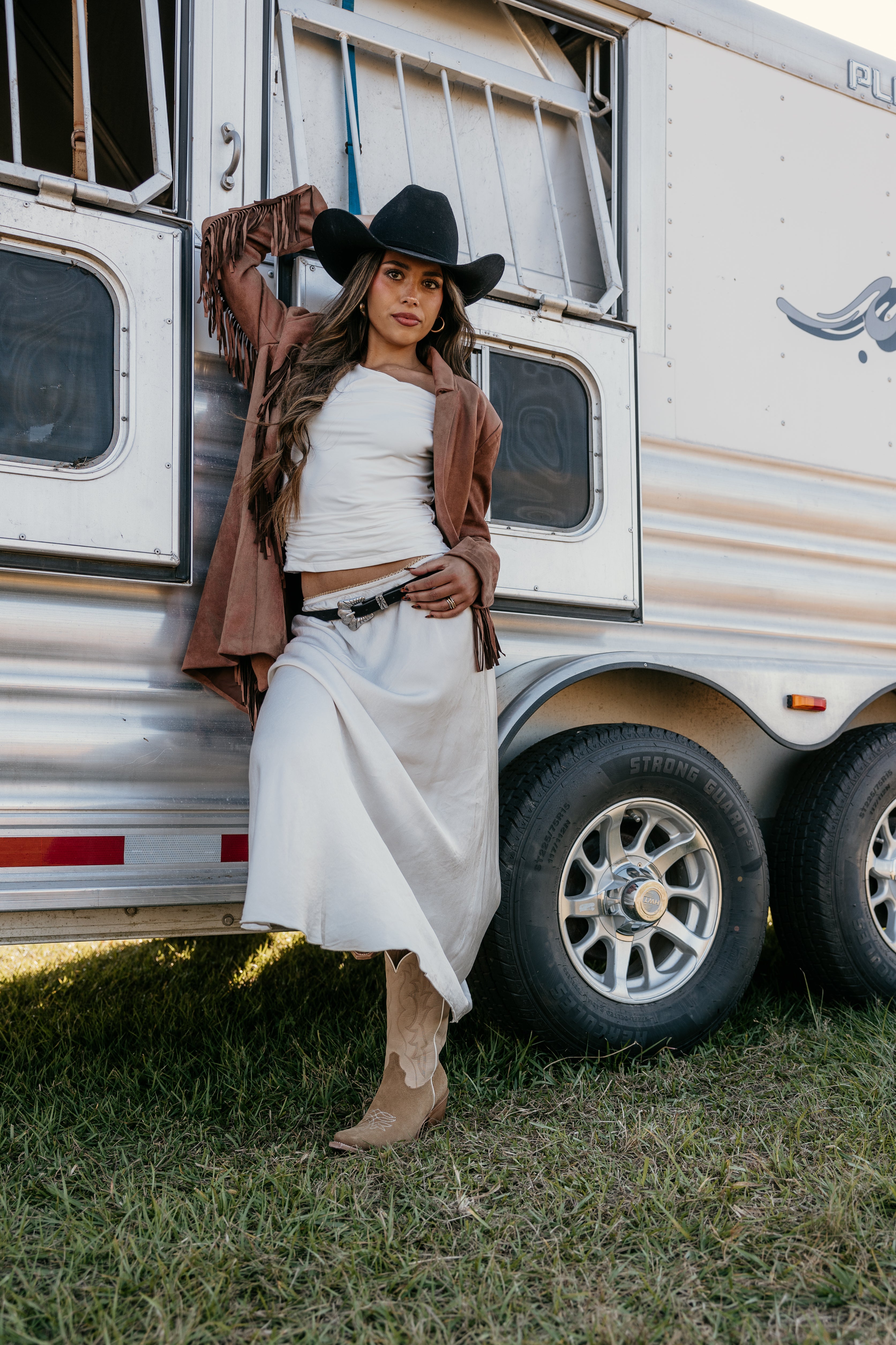 Woman in western boho outfit with suede fringe jacket, white skirt, and cowboy hat by horse trailer