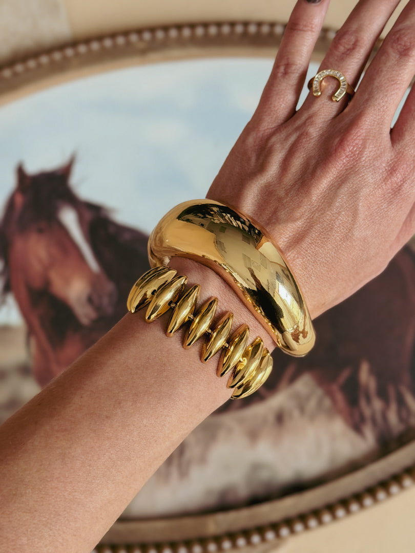 Gold western jewelry, including chunky bracelets and a horseshoe ring, worn on a hand with a horse painting in the background.