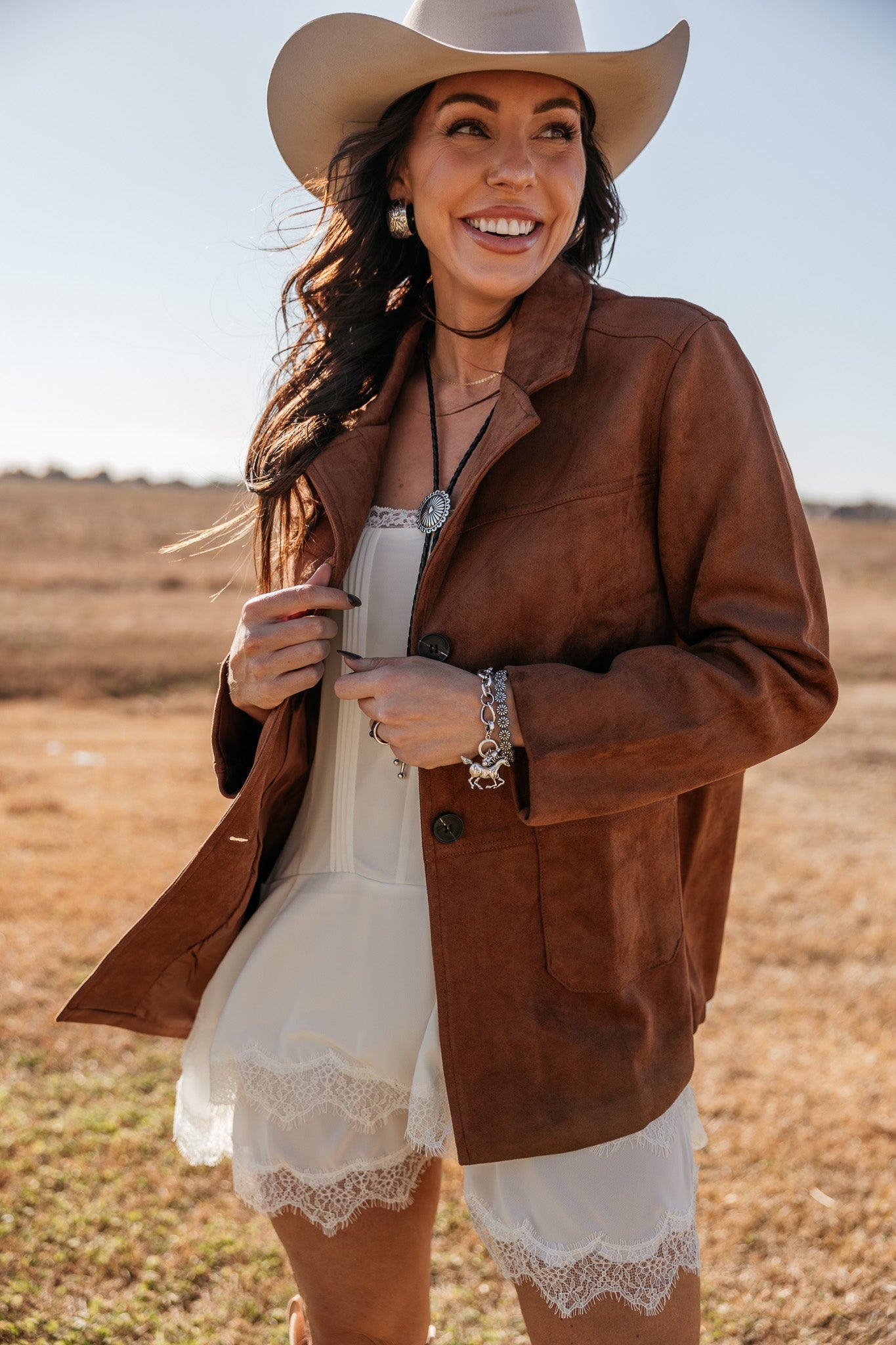 Woman in cowgirl hat, brown suede jacket, and white lace dress in western outdoor setting