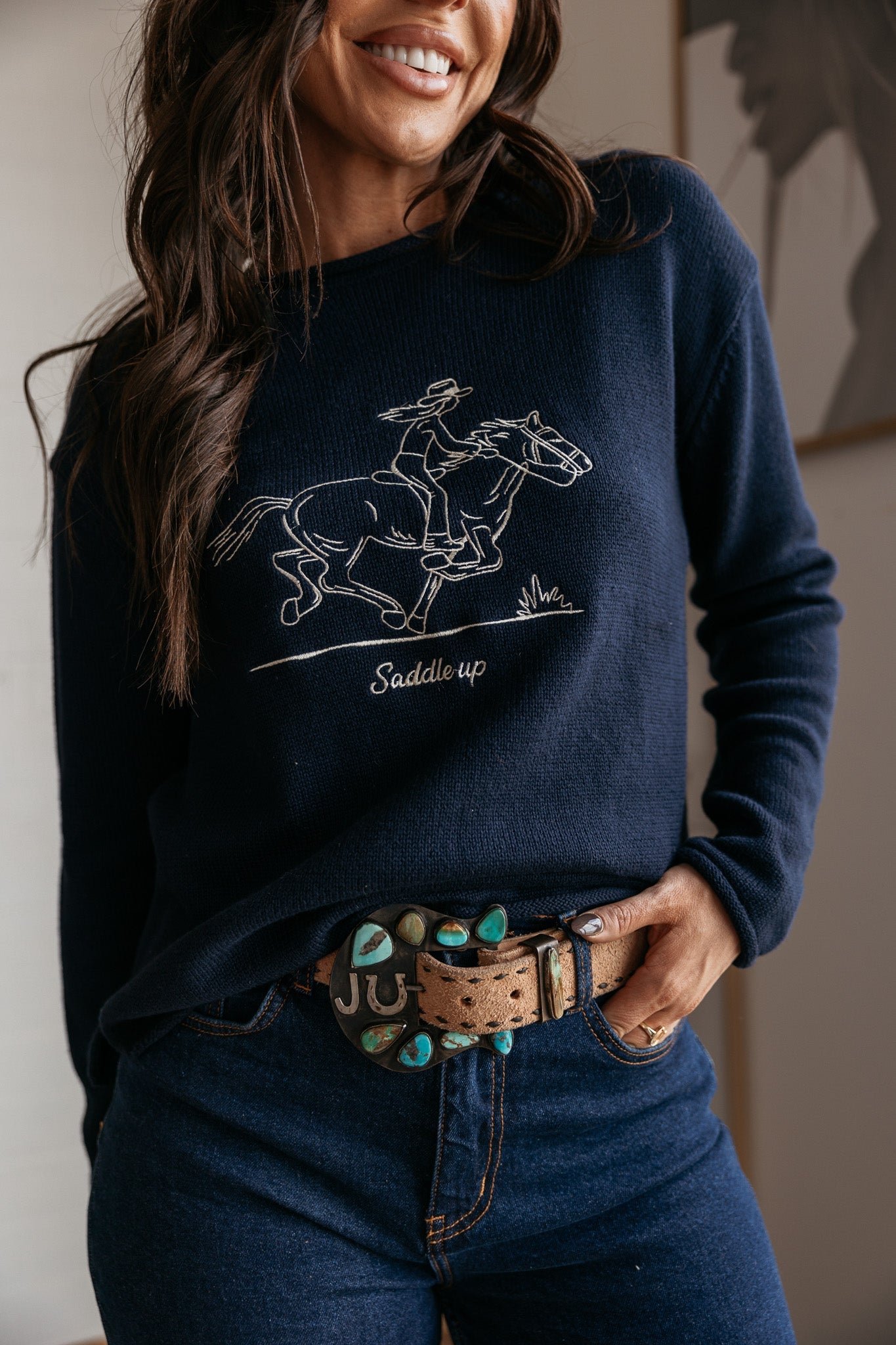 Woman in navy western sweater with cowboy and horse graphic, turquoise concho belt and jeans