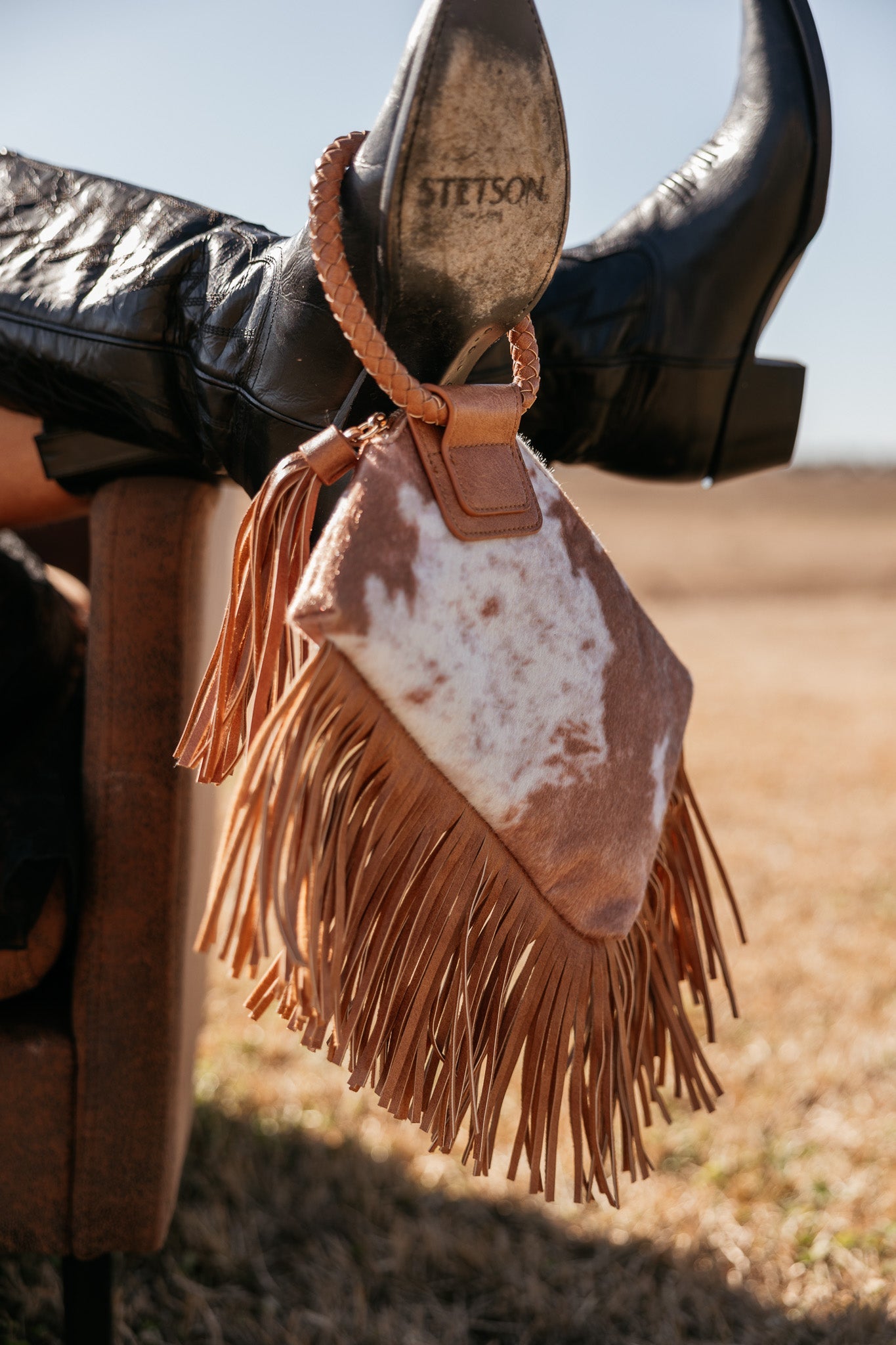 Western cowhide fringe clutch bag with braided handle, black Stetson boots, outdoors