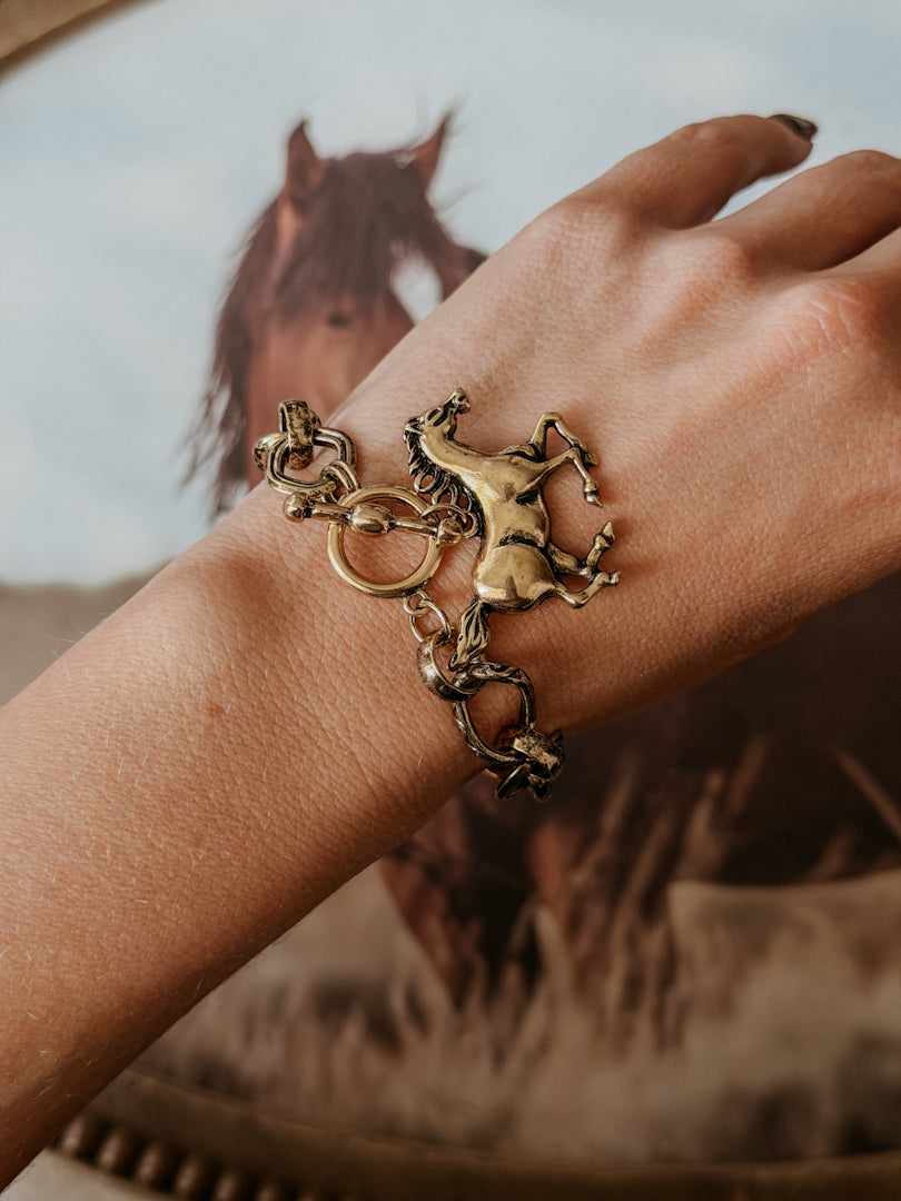 Western horse charm bracelet on wrist with galloping horse artwork in background