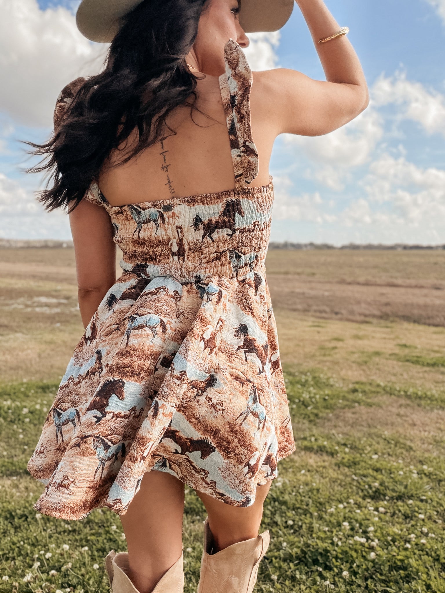 Woman in a patterned dress and cowboy boots standing in an open field with a blue sky.