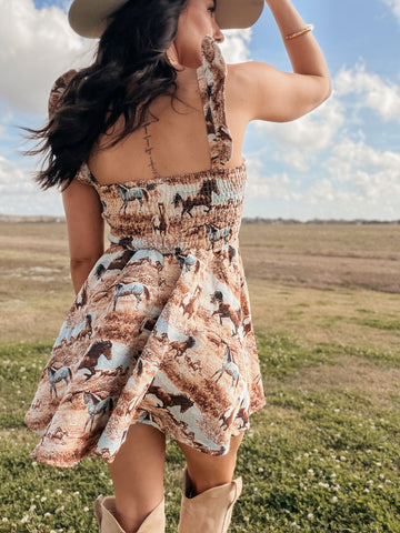 Woman in a patterned dress and cowboy boots standing in an open field with a blue sky.