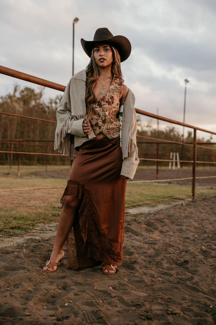 Woman in western boho outfit with cowgirl hat, fringe jacket, and brown maxi skirt outdoors