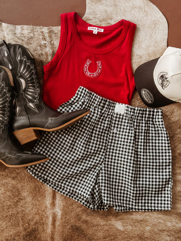 outfit featuring black embroidered cowboy boots, a red ribbed tank top with a white horseshoe graphic, black and white gingham shorts with an elastic waistband, and a two-tone trucker hat with a horse and horseshoe graphic.